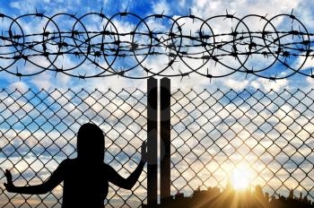 The concept of the refugees. Silhouette of a refugee near the fence with barbed wire against the evening sky and the city in the distance