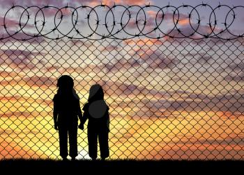 Concept of the refugees. Silhouette of hungry children in desperate refugees near the fence at sunset