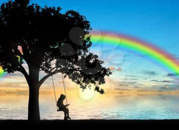 Silhouette of a little girl reading a book sitting on a swing by the sea with a rainbow. Conceptual image of childhood