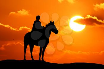Silhouette or woman and horse running across horizon as the sun goes down.