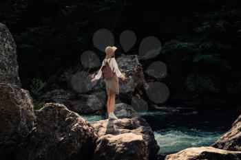 woman traveler with backpack and hat walking in amazing mountains and forest near river with deep blue water