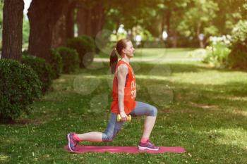 Sport training concept. The girl is engaged in fitness in nature. Side view of young woman doing stretching exercise using dumbbells. Active athlete working out on fitness routine to lose weight.