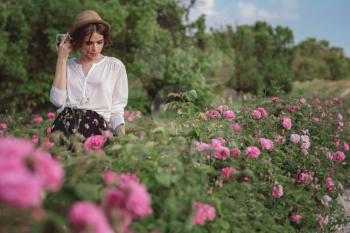 Beautiful young woman with curly hair posing near roses in a garden. The concept of perfume advertising