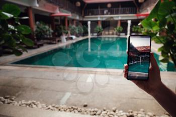 hand holding phone on background of the pool in hotel. photo camera on the screen. close up hand hold smartphone take a picture of summer vacation.