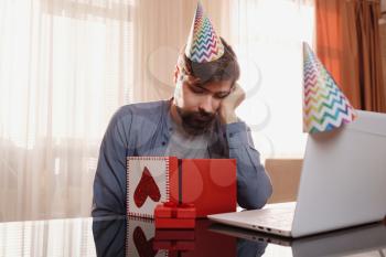 Upset young man sitting at the table and opennig the gift box. Concept of solitude in quarantine during the Coronavirus Pandemic COVID-19. A bad gift is a frustrated birthday man.