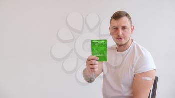 Adult man wearing t-shirt holding green International Certificate of Vaccination on white background. Traveling Immune passport, as proof vaccinated against Covid-19.