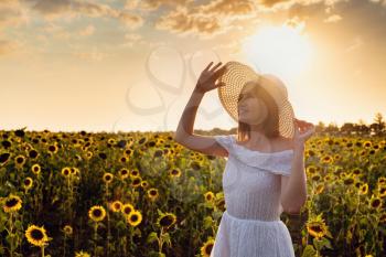 Beautiful young girl enjoying nature on the field of sunflowers at sunset. Asian girl in a cute white dress and hat enjoys summer and vacation.