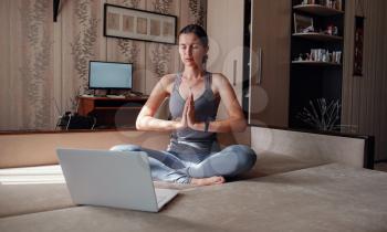 Time for yoga. Attractive young woman exercising and sitting in yoga lotus position while resting at home. View of a woman conducting virtual fitness class at home on a video conference.