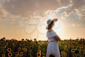 Beautiful young woman enjoying nature on the field of sunflowers at sunset. Asian woman in a cute white dress and hat enjoys summer and vacation.