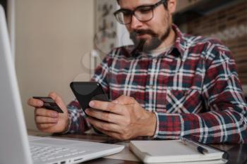 Handsome man using credit card to pay online with smartphone. Shopping Online, Banking system. guy in a plaid shirt sitting in his home kitchen