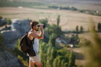 Guy looking at binoculars in hill. man in t-shirt with backpack. Young Caucasian man during hike in valley landscape