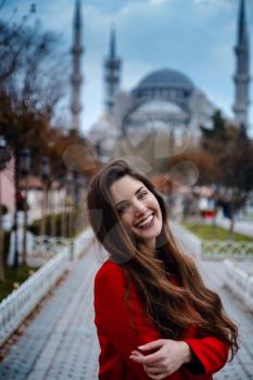 Latin American woman or Turkish woman in a red stylish coat in front of the famous blue mosque in Istanbul, Photo of traveler on the background of a mosque on autumn day.