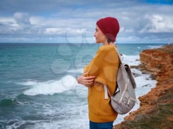 Hipster woman traveler with backpack in yellow sweater and red hat enjoying wind on the coast. storm and thunderstorms in the distance over the sea, the happiness and freedom of travel and discovery