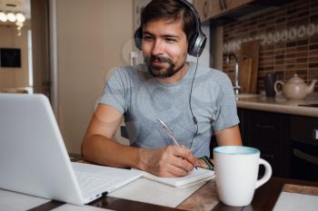 Focused young man wear headphones study online watching webinar podcast on laptop listening learning education course conference calling make notes sit at work desk, elearning concept