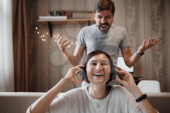 angry husband shouting at wife sitting on sofa and covering ears with headphones. the woman ignores the man