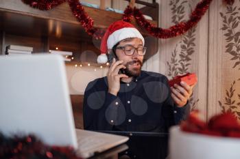 Close up portrait of joyful Caucasian handsome man in santa hat sitting in cozy room xmas decoration calling by smartphone with happy face . New Year. Merry Christmas