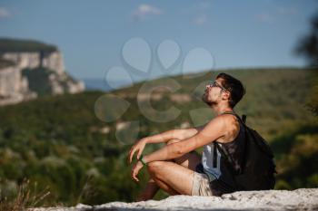 Young male hiker with backpack relaxing on top of a mountain during calm summer sunset - scenery from vacation. Tired traveling man resting on hill.