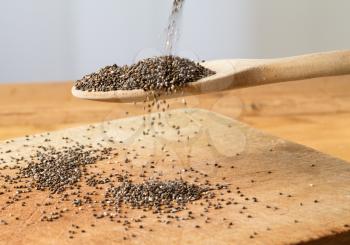 Superfood known as black chia seeds are poured into a wooden backing spoon over wood bread board on kitchen table
