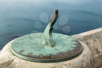 Green stained copper or brass sundial fixed to stone wall of castle overlooking the ocean far below