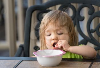 Small caucasian baby girl eating with fingers from bowl on outdoor table