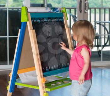 Young two year old girl drawing with finger on blackboard on an easel during playtime