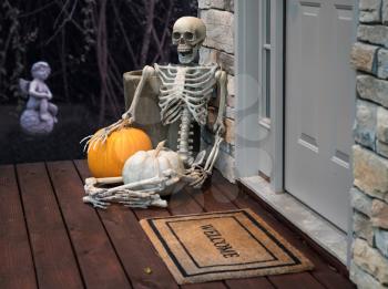 Scary skeleton sitting on porch of modern house by front door with welcome mat and garden in background. Funny Halloween image for trick or treat
