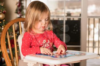 Young female baby girl in high chair making a jigsaw on a plastic tray at Christmas with tree in background