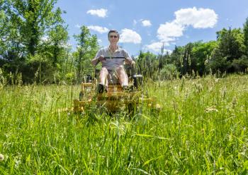 Senior retired male cutting very deep grass in a meadow or field after leaving it to grow for far too long before cutting using yellow zero-turn lawn mower