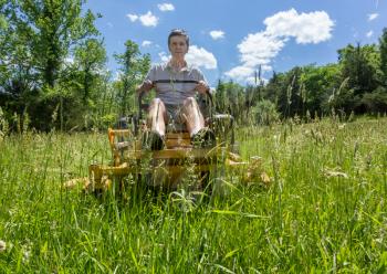 Senior retired male cutting very deep grass in a meadow or field after leaving it to grow for far too long before cutting using yellow zero-turn lawn mower