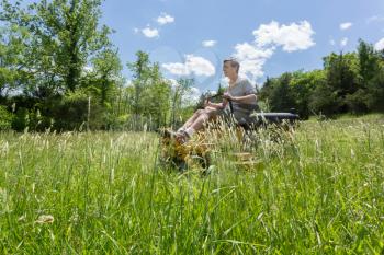Senior retired male cutting very deep grass in a meadow or field after leaving it to grow for far too long before cutting. He is mowing sideways to the camera using yellow zero-turn lawn mower