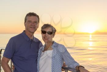 Middle aged couple on an ocean sunset cruise and leaning against the railings. Smiling and facing the camera.