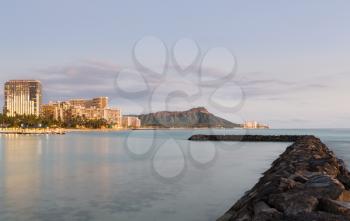 Panorama of the skyline of Waikiki at dusk taken with a long exposure to blur out movement in the water. Breakwater leads the eye towards the volcano of Diamond Head in Hawaii