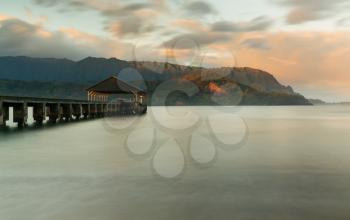 Rising sun illuminates the peaks of Na Pali mountains over the calm bay and Hanalei Pier in long exposure photo