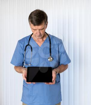 Senior male caucasian doctor with stethoscope in medical scrubs looking down and holding electronic tablet for message