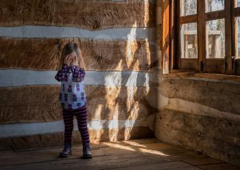 Small and lonely caucasian baby girl or toddler standing inside empty old wooden cabin to suggest poverty or recession