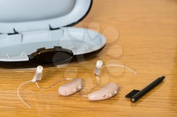 Macro close up of a matched pair of tiny modern hearing aids with case and brush on wooden bedside dressing table