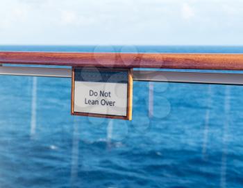 Warning sign Do Not Lean Over on the teak railing of cruise ship above the ocean