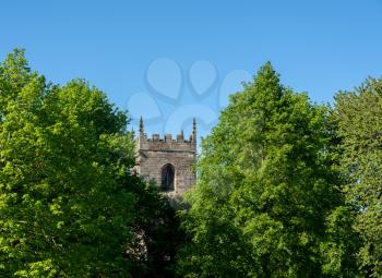 Idyllic british scene of stone church tower appearing between the leaves of two large spring trees against blue sky
