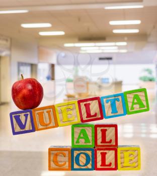 Vuelta al Cole translates to Back to School in spanish with wooden blocks with red apple on top. Background is a school entrance or corridor