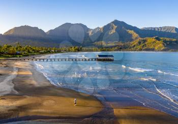 Aerial image at sunrise off the coast over Hanalei Bay and pier on Hawaiian island of Kauai with a man standing alone on the beach