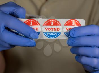Hands in medical gloves holding roll of I Voted Today buttons ready for in person voters in Presidential election