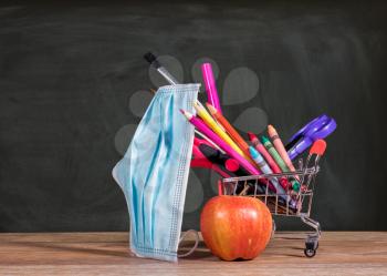 Back to school during coronavirus pandemic with pencils, crayons and face mask in shopping cart with apple against chalkboard