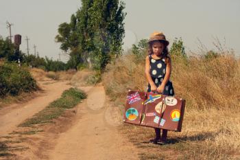 Little girl with suitcase in which the stickers from various countries waiting at the road passing trucks to get hitchhiking