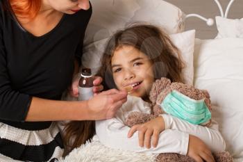Smiling little girl with a favorite teddy bear on whom they put on a gauze bandage is lying sick in bed and her mother is drinking her medicine.