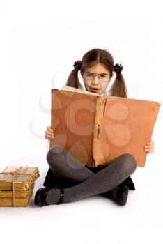 A little schoolgirl in glasses holds an enormous old book in her hands and reads it sitting on the floor