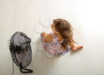The little girl is sitting in front of the fan and is cooling. Air streams her hair