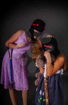 Two hooligans of a teenager girl in bright elegant dresses, but in gangster hats, balaclava with heavy clubs, one of them comforts a crying second girl
