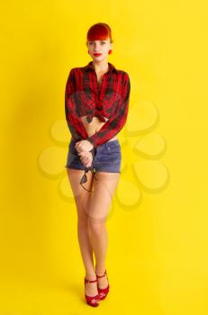 The girl in retro style in a bright checkered shirt knotted on her stomach and shorts posing on a bright yellow background