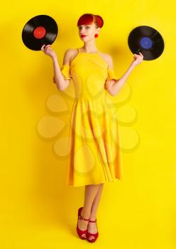 The girl in retro style, in a yellow dress stands with two vinyl records on a bright background