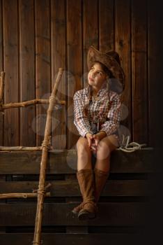 Little girl in a wide-brimmed cowboy hat and traditional dress in high boots and with a lasso posing on a dark wooden background